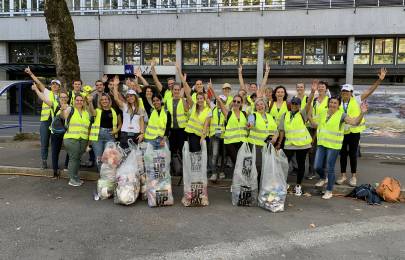 Bild: Volunteers 2023. Eine Gruppe von Freiwilligen in gelben Warnwesten steht vor einem Gebäude, hebt die Hände und posiert mit mehreren grossen Müllsäcken nach einer gemeinsamen Aufräumaktion.