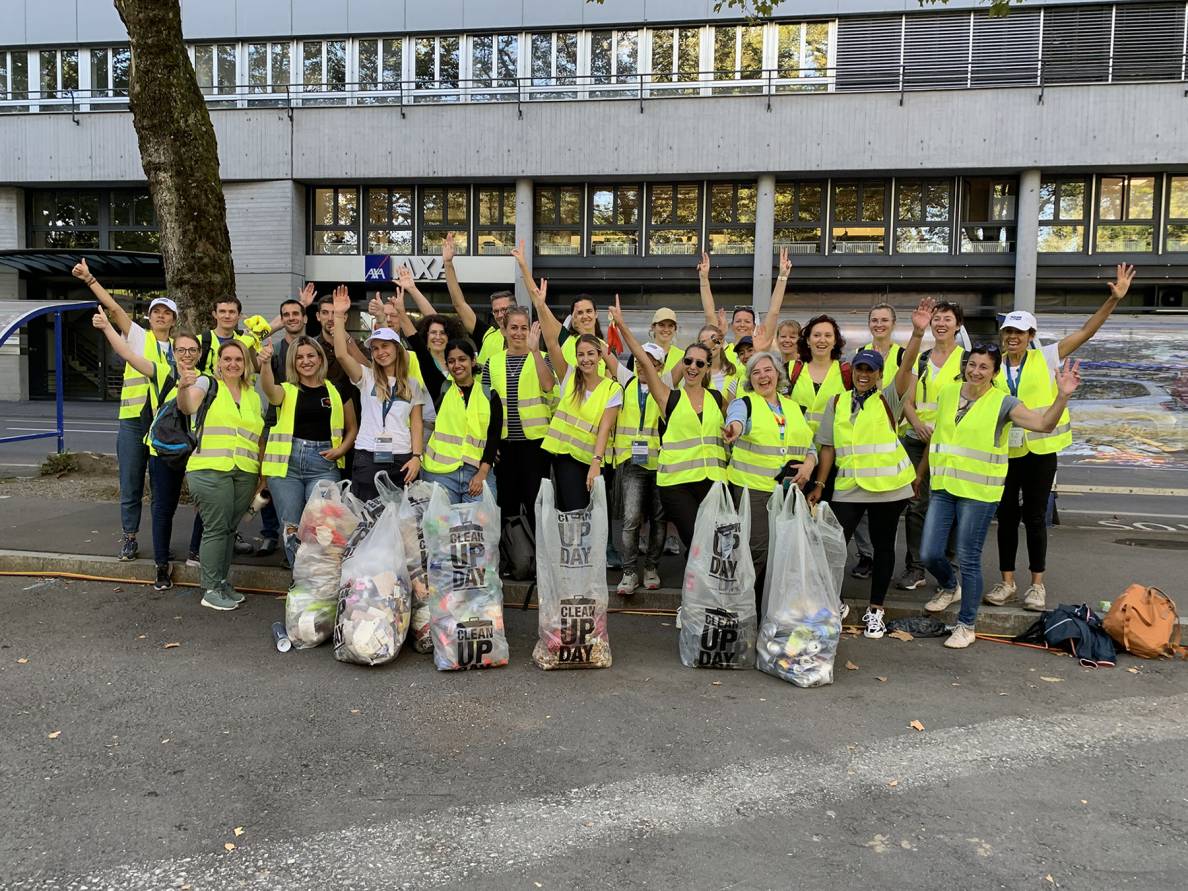 Bild: Volunteers 2023. Eine Gruppe von Freiwilligen in gelben Warnwesten steht vor einem Gebäude, hebt die Hände und posiert mit mehreren grossen Müllsäcken nach einer gemeinsamen Aufräumaktion.