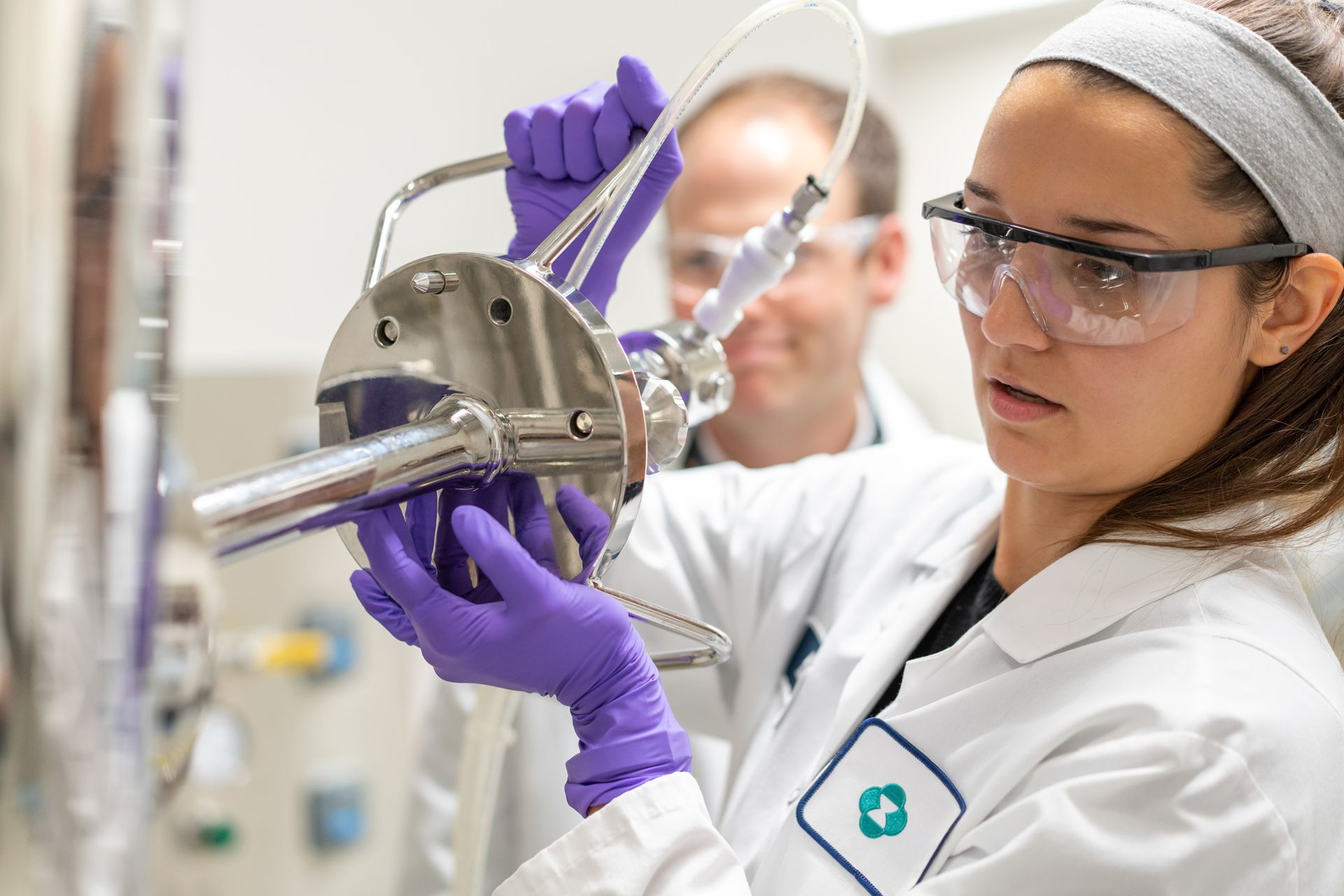 Eine Wissenschaftlerin mit Laborkittel, Schutzbrille und violetten Handschuhen justiert sorgfältig ein Laborgerät mit Schläuchen, während ein Kollege im Hintergrund zuschaut.