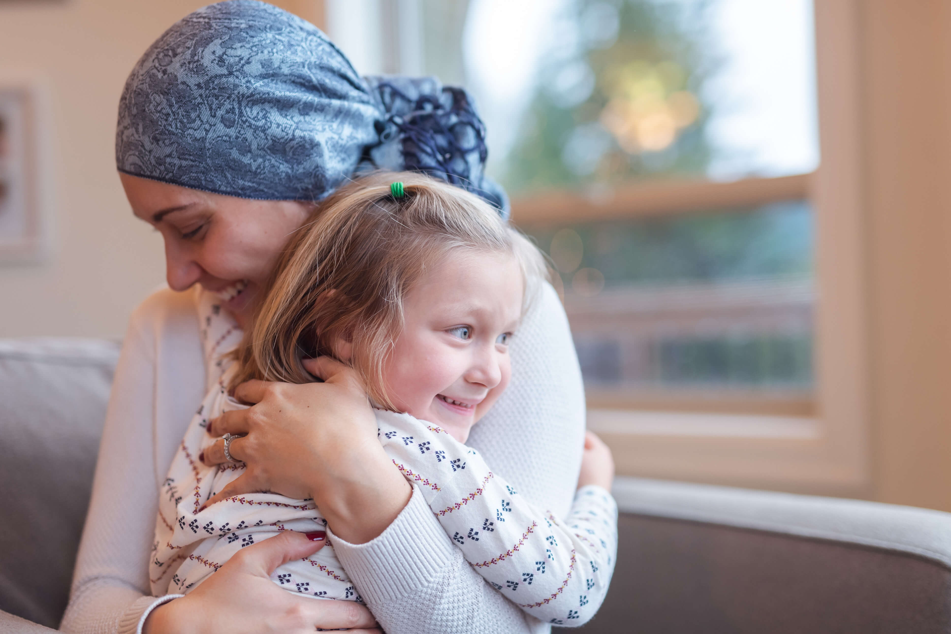 A mother wearing a headscarf hugs her smiling child on the sofa. She radiates warmth and closeness.