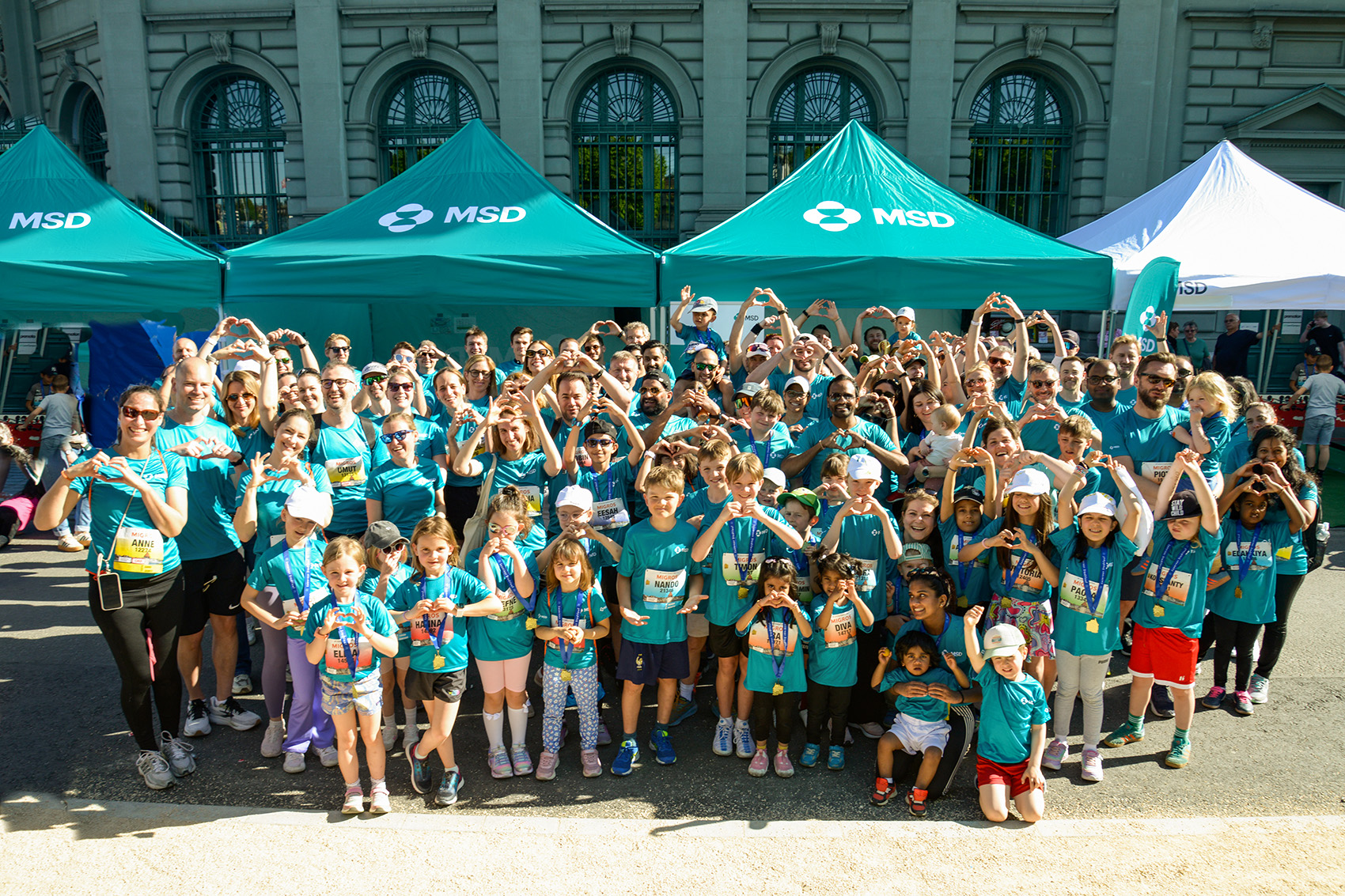 Large group of adults and children in turquoise MSD T-shirts, posing in front of green MSD tents and forming heart shapes with their hands at a running event