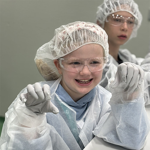 Photo: Enfant en tenue de laboratoire souriant et levant les mains gantées à une table de labo.
