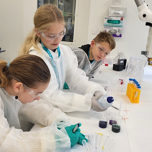 Photo: Trois enfants en tenue de laboratoire travaillent ensemble avec des pipettes et des liquides colorés.