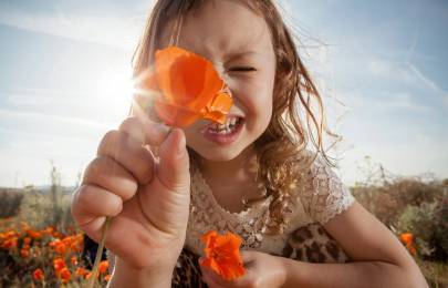 Une enfant souriante en plein air tient un coquelicot orange près de la caméra, couvrant partiellement son visage; à l’arrière-plan, on voit d’autres fleurs orange et un ciel lumineux.