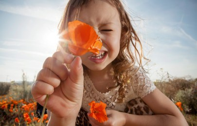 Une enfant souriante en plein air tient un coquelicot orange près de la caméra, couvrant partiellement son visage; à l’arrière-plan, on voit d’autres fleurs orange et un ciel lumineux.