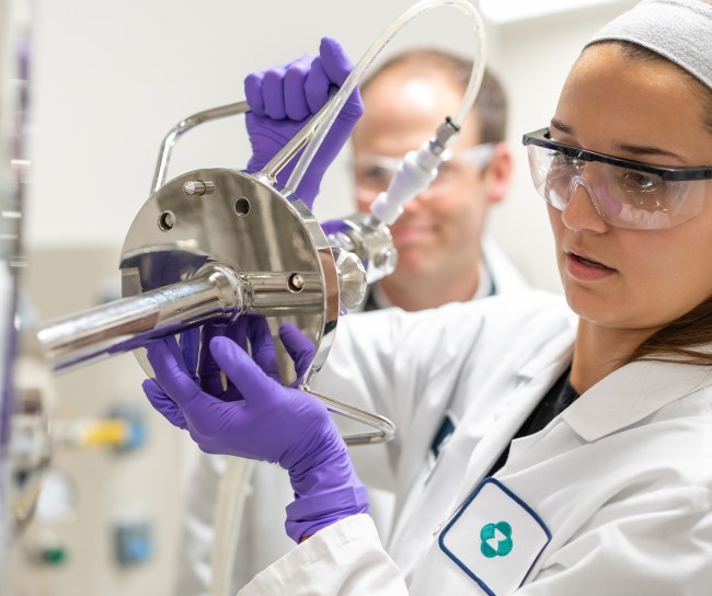 Une scientifique portant une blouse de laboratoire, des lunettes de protection et des gants violets ajuste soigneusement un équipement de laboratoire avec des tuyaux, tandis qu’un(e) collègue observe à l’arrière-plan.